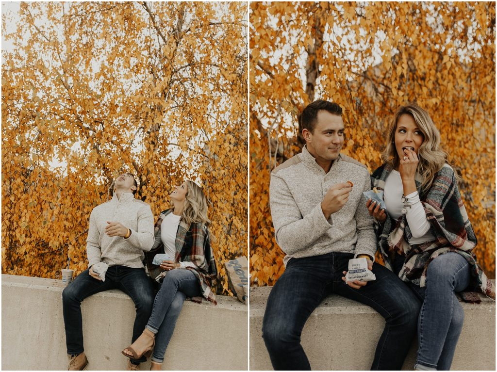 engaged couple sitting on a wall in front of an orange fall tree eating a burger and fries