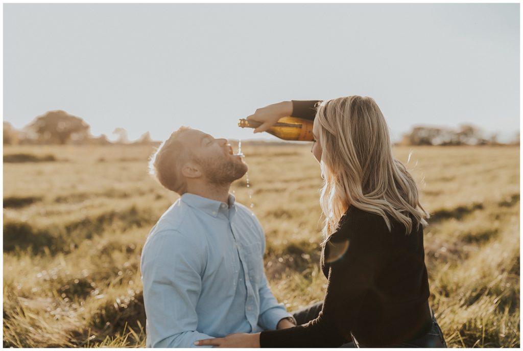 couple taking engagement photos and popping champagne in a field during golden hour
