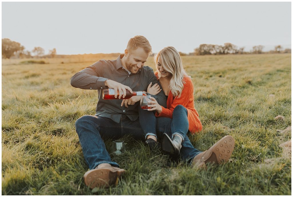 couple taking engagement photos and sipping wine in a field during golden hour